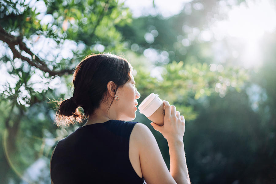 Low angle view of young Asian woman relaxing in a park, drinking a cup of coffee while surrounded by beautiful nature and the warmth of sunlight Photograph by D3sign