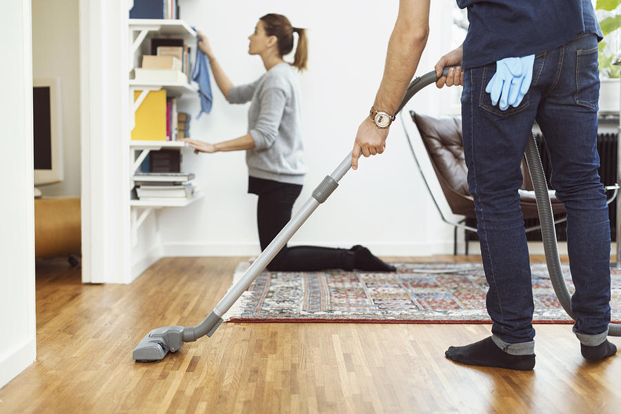 Low section of man vacuuming floor while woman cleaning shelves in background at home Photograph by Maskot