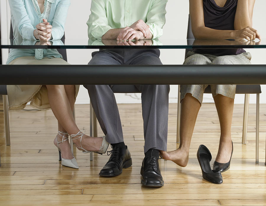 Low Section View of Two Businesswomen Touching the Feet of a Businessman Under a Table Photograph by Digital Vision.