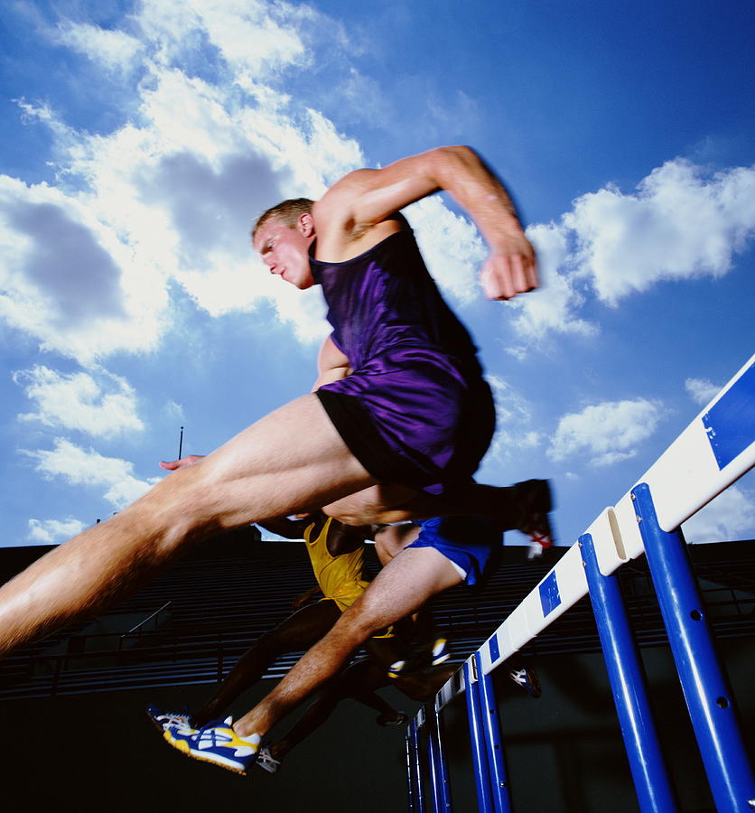 Male athletes leaping hurdles in track and field race, low angle. Photograph by White Packert