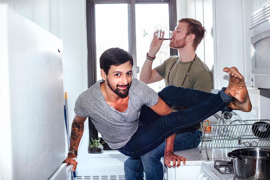 Male couple at home, mid adult man drinking whilst his partner balances on kitchen appliances Photograph by Ashley Corbin-Teich