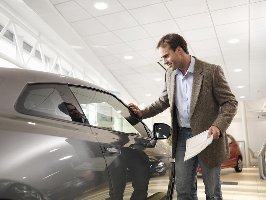 Male customer holding paper, looking at car with hand touching car in car dealership Photograph by Monty Rakusen