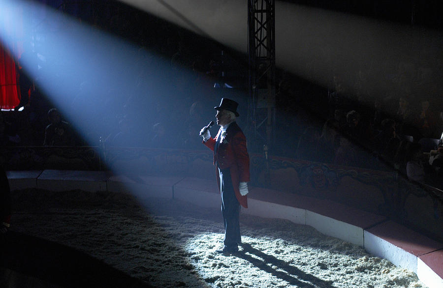 Male Ringmaster Stands in a Spot lit Circus Ring, Making an Announcement to the Audience Photograph by Adrian Peacock