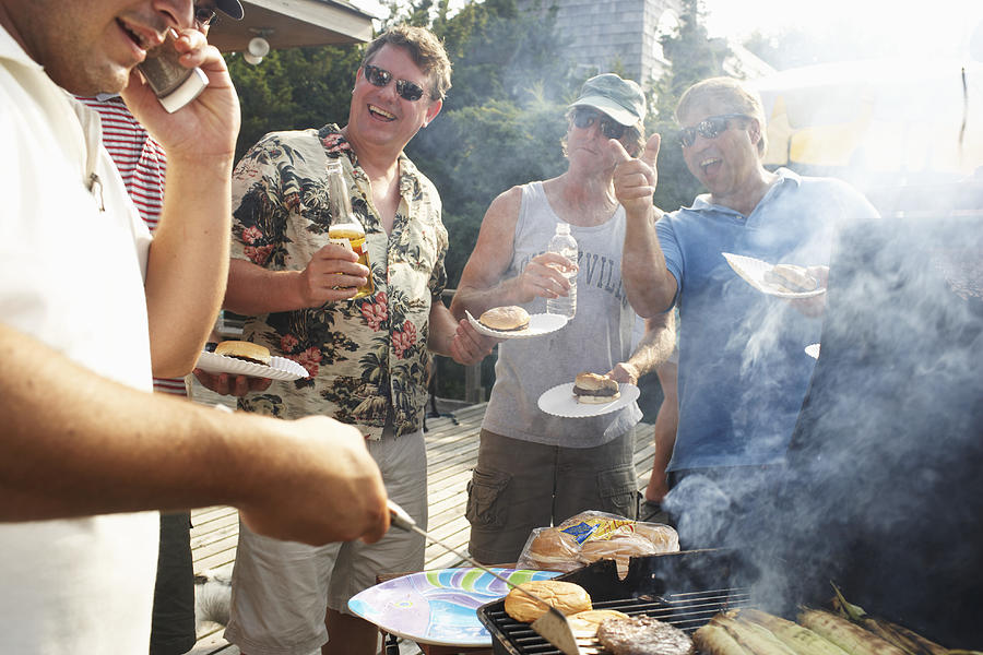 Man barbecuing using mobile phone, friends laughing in background Photograph by Christopher Robbins