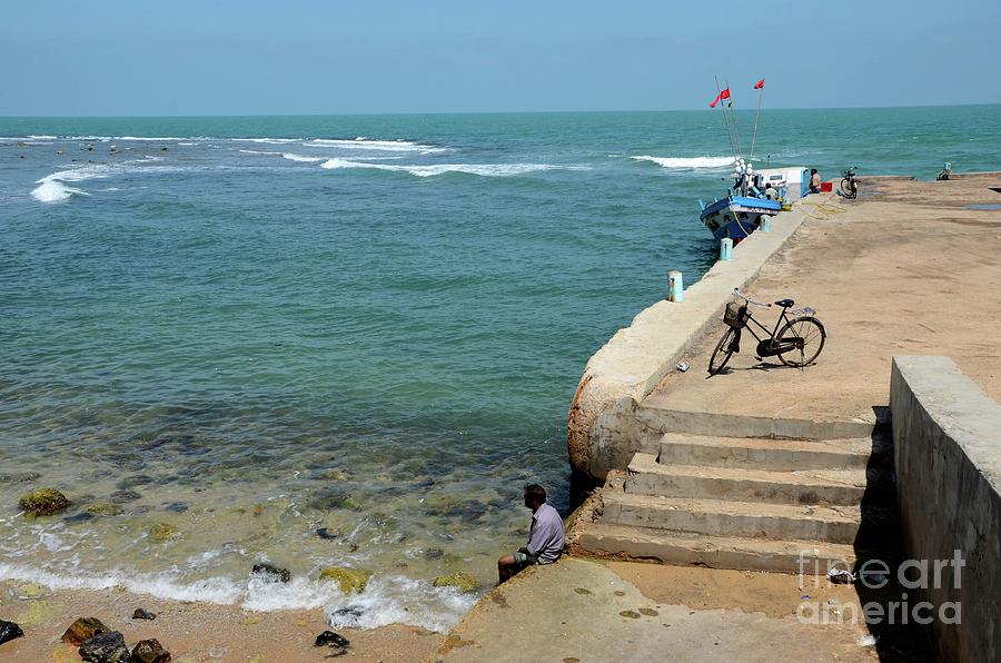 Man dips feet in water at beach pier with fishing vessel  near steps in Jaffna Sri Lanka Photograph by Imran Ahmed