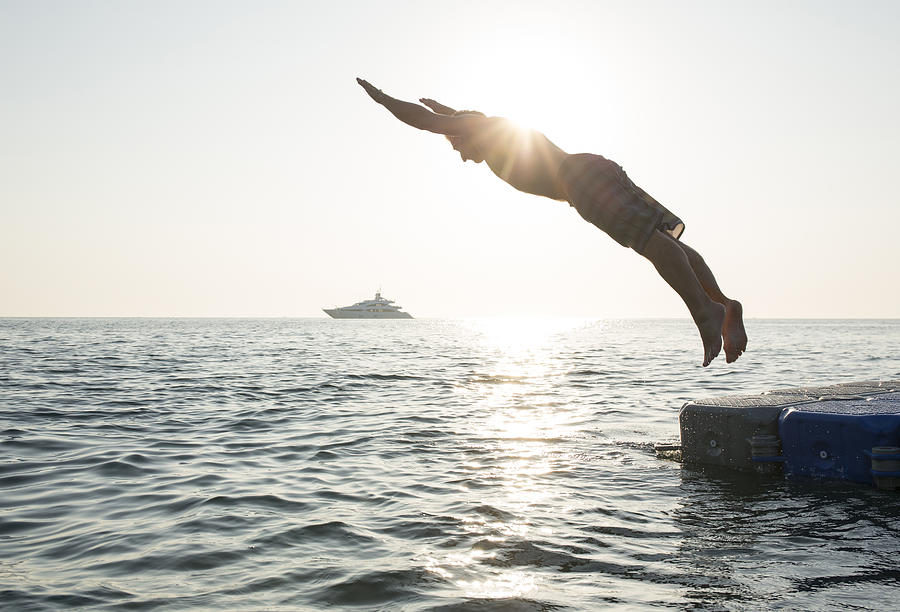 Man dives into tranquil sea, from pier, boat Photograph by AscentXmedia