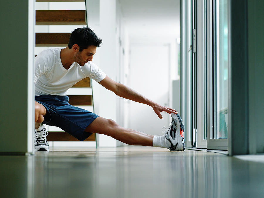 Man in sports clothes performing leg stretch in hallway, ground view Photograph by Michael Blann