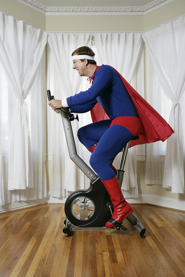 Man in superhero costume on exercise bike, side view Photograph by Lauren Nicole