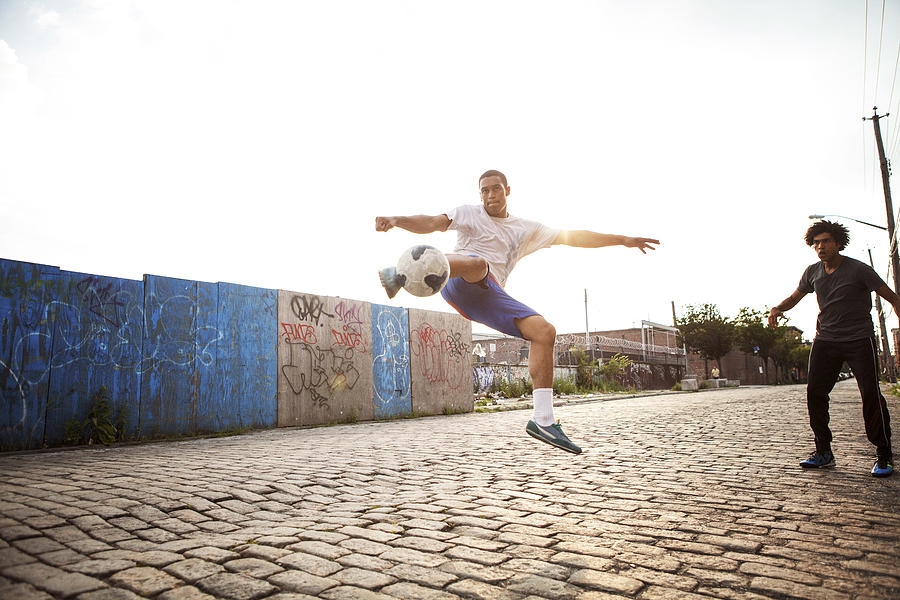 Man kicking soccer ball while friend looking at street Photograph by Cavan Images