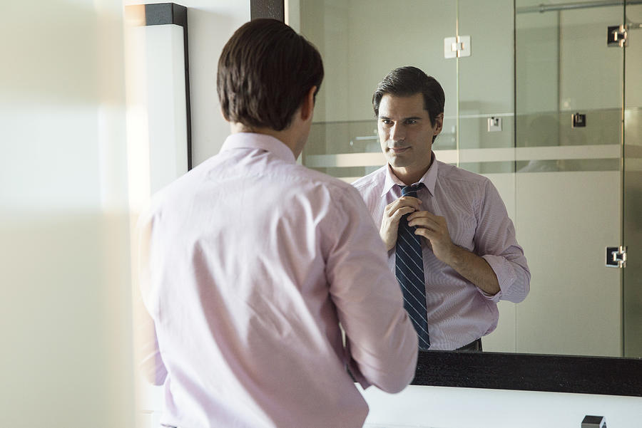 Man looking in bathroom mirror, adjusting necktie Photograph by PhotoAlto/Sigrid Olsson