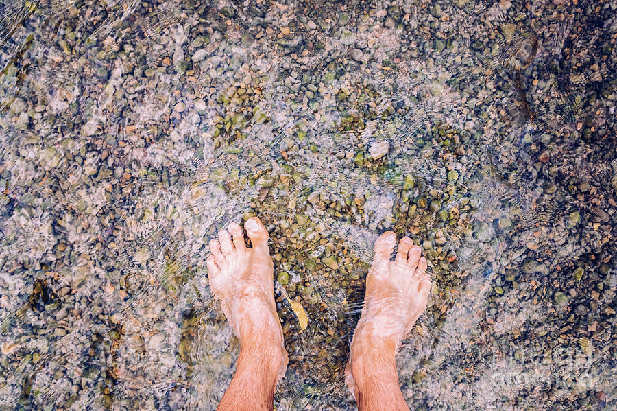 Man looks at his feet submerged in the water of a river on the pebbles. Photograph by Joaquin Corbalan