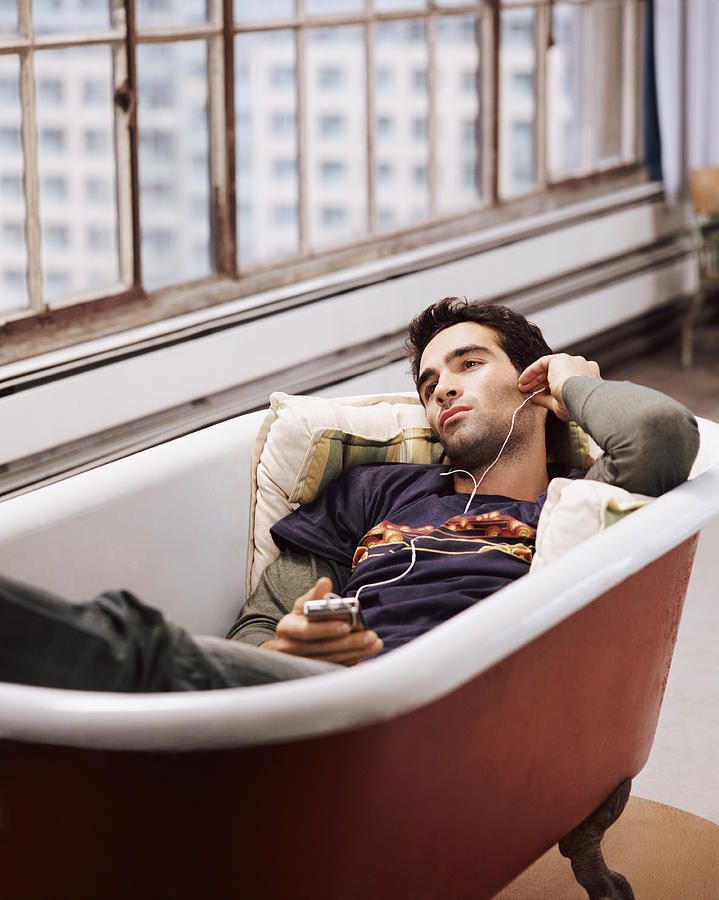 Man Lying in an Empty Bath Listening to Music on an MP3 Player Photograph by Digital Vision.