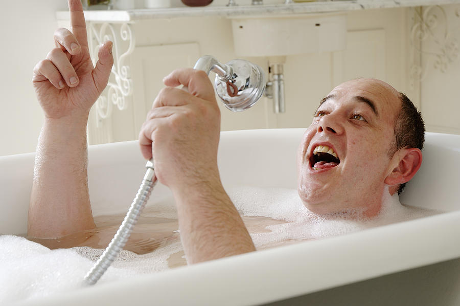 Man lying in bath singing into shower head, close-up Photograph by James Darell