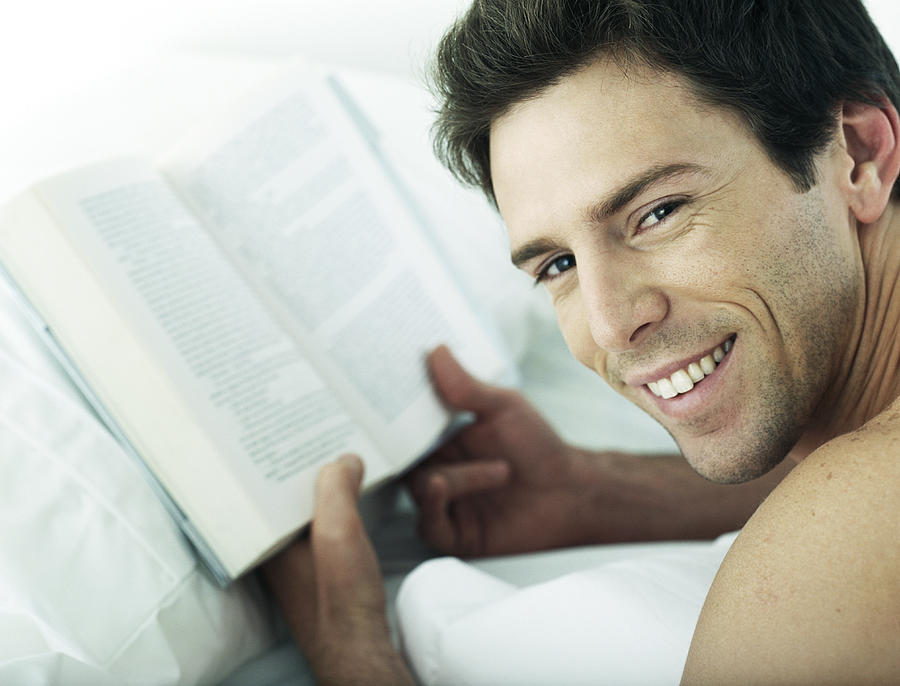Man lying on stomach holding book, looking over shoulder at camera, close-up Photograph by Laurence Mouton