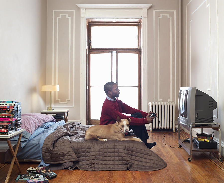 Man playing video game in flat, dog on bed Photograph by Mark Lund