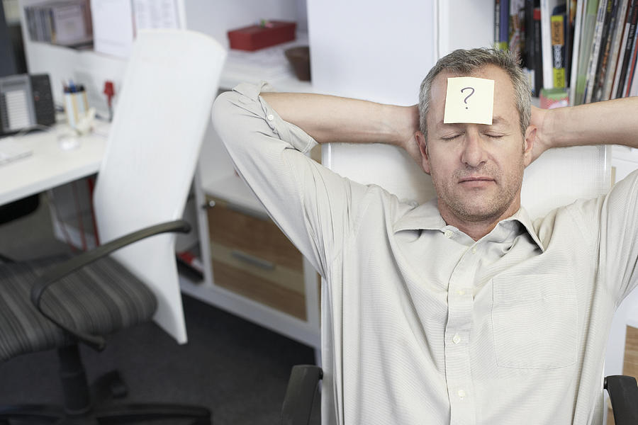 Man relaxing in office with question mark written on adhesive note stuck to forehead Photograph by David Lees