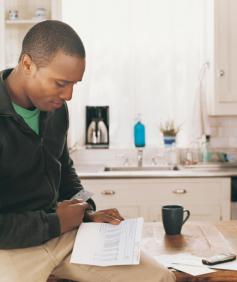 Man Sitting in a Kitchen Looking at a Bank Statement Photograph by Digital Vision.