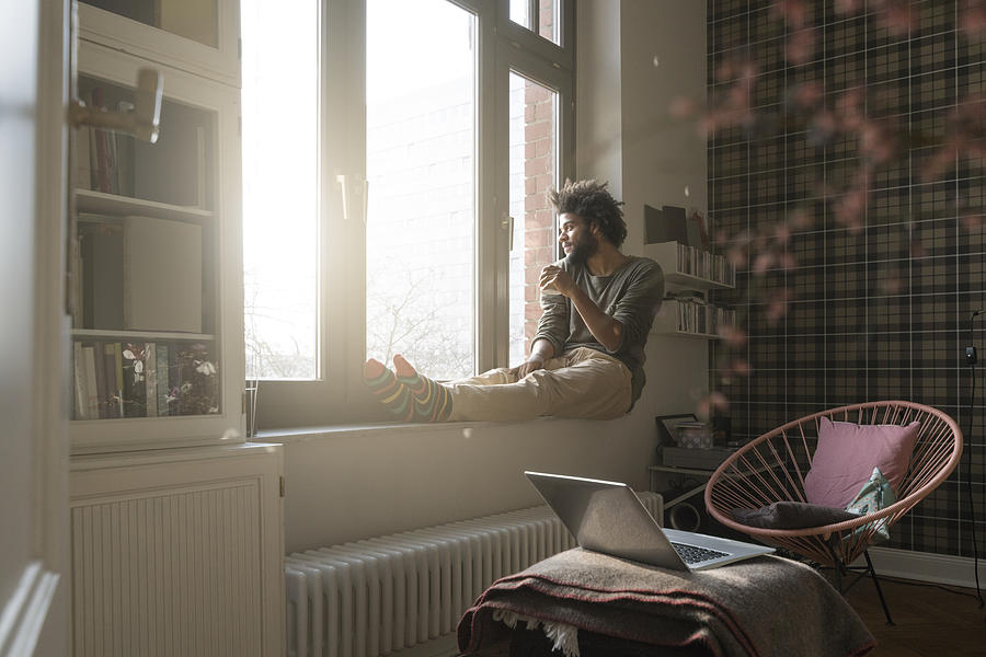 Man sitting on window sill in living room looking outside holding a cup Photograph by Westend61