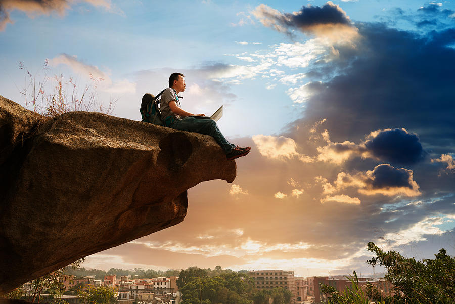 Man standing at the edge of a cliff using laptop Photograph by Baona