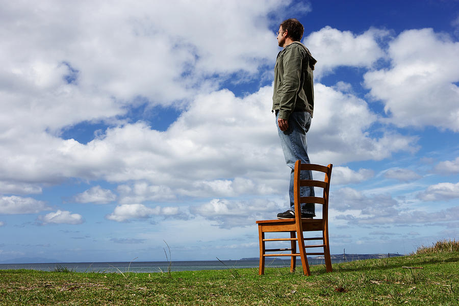 Man standing on chair in field, looking at landscape, side view Photograph by Paul Sutherland