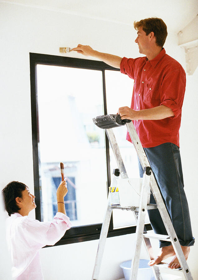 Man standing on ladder, painting wall, woman holding up brush Photograph by John Dowland