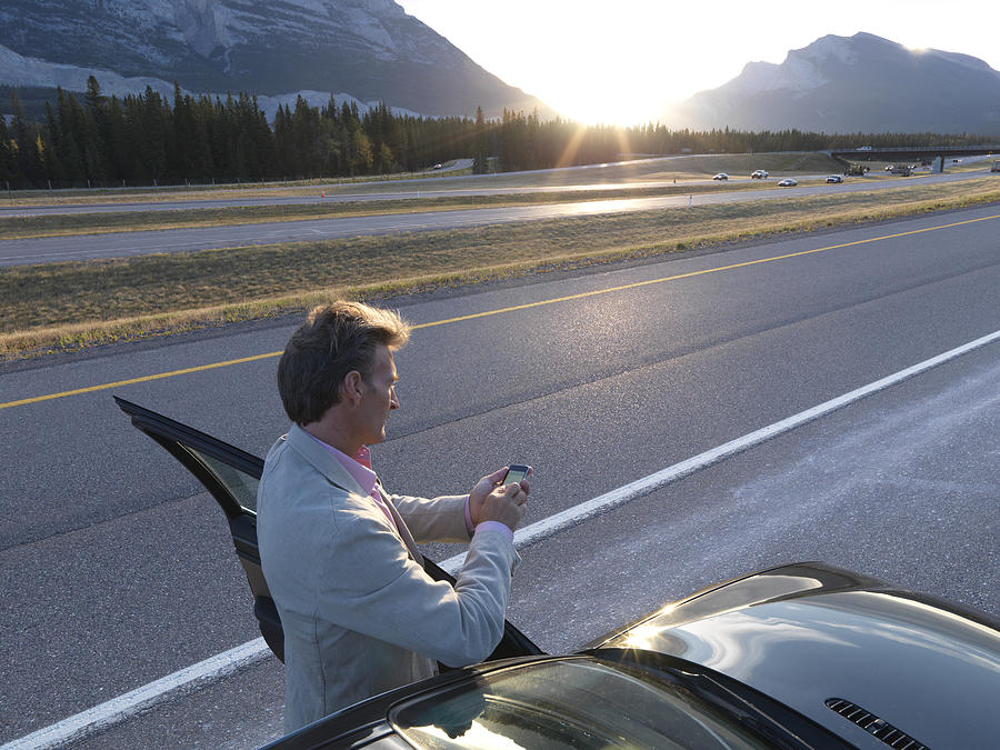 Man text messages besides car at edge of mtn road Photograph by Ascent/PKS Media Inc.