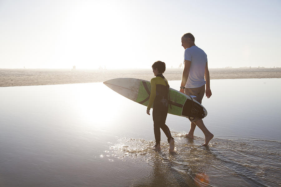 Man walking with grandson carrying surf board Photograph by Alistair Berg