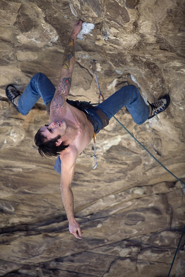 Man wearing blue jeans, without shirt, with tattoos hangs upside down from one arm while climbing an overhang. Photograph by Aaron Black