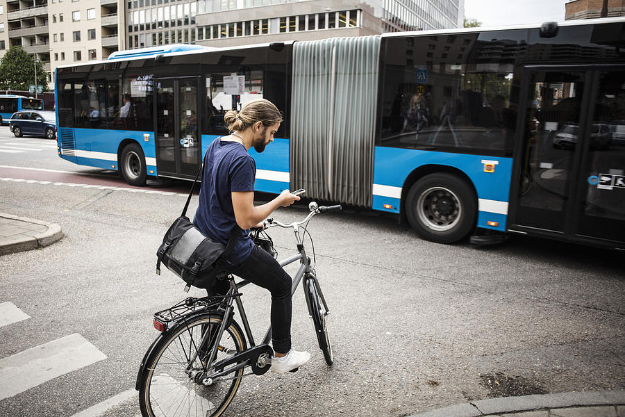Man with bicycle using mobile phone while standing on city street against articulated bus Photograph by Maskot
