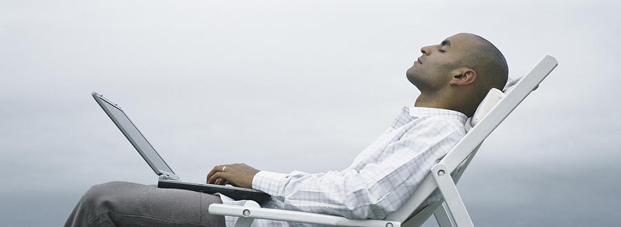 Man with eyes closed in lounge chair with laptop on lap, sky in background, side view Photograph by Matthieu Spohn