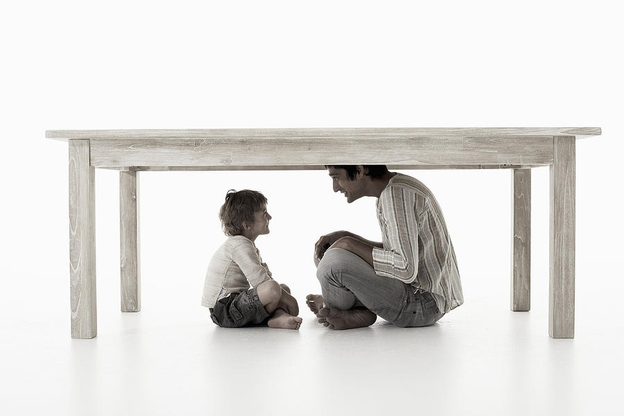 Man with son (4-5 years) sitting under table, studio shot Photograph by Knowlesie