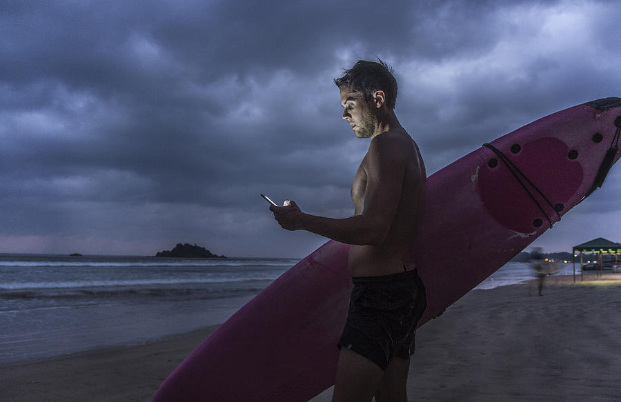 Man with surfboard using smart phone at dusk Photograph by David Trood