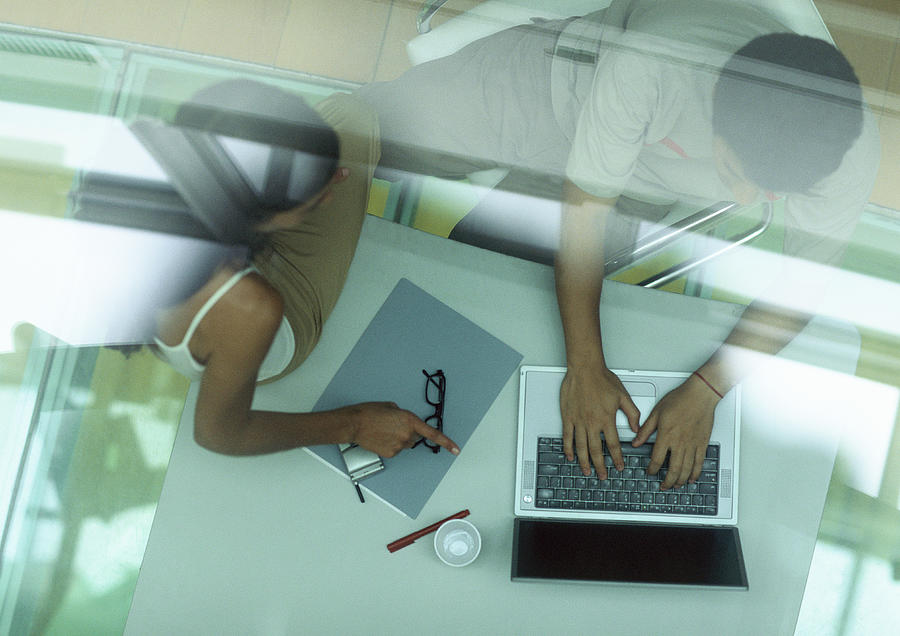 Man working on computer, woman sitting on table pointing at computer, behind reflection on glass, high angle view. Photograph by Gerard Launet
