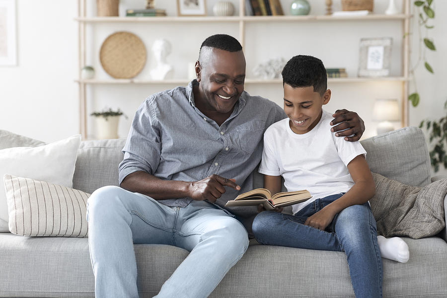 Mature african american man reading book with his grandson at home Photograph by Prostock-Studio