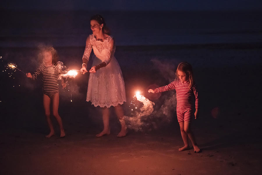 Mature bride lighting bengal fire with daughters on the beach at dusk. Photograph by Martinedoucet