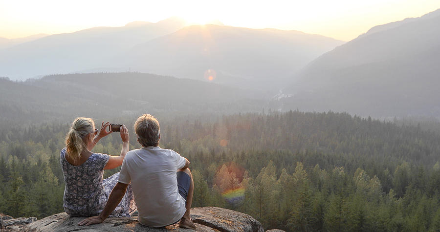 Mature couple relax on mountain ledge, look out to view Photograph by AscentXmedia