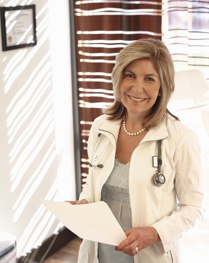 Mature female doctor holding medical chart, smiling, portrait Photograph by Ascent/PKS Media Inc.