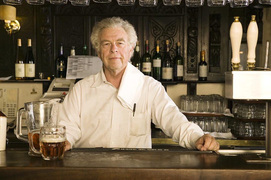 Mature male bartender standing behind bar, smiling, portrait Photograph by Caroline Schiff