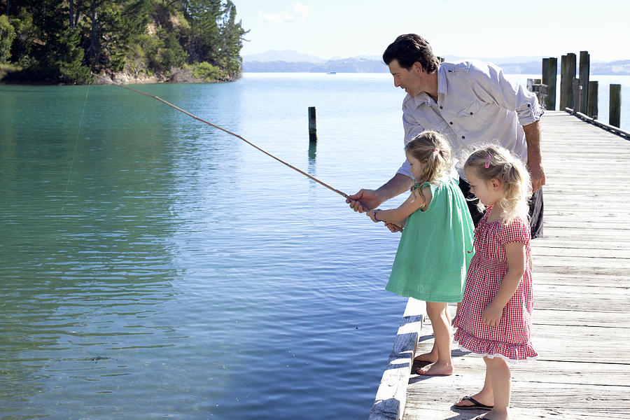 Mature man and two young girls fishing from pier, New Zealand Photograph by Axel Bernstorff
