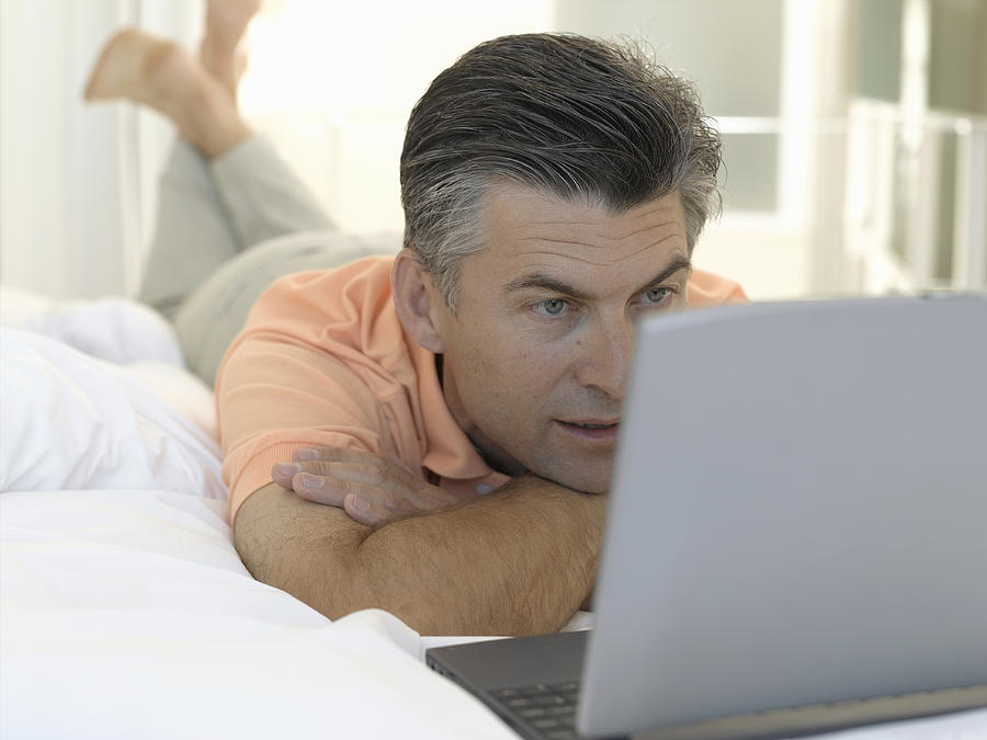 Mature man lying on bed looking at laptop computer, close-up Photograph by Matthias Heitmann