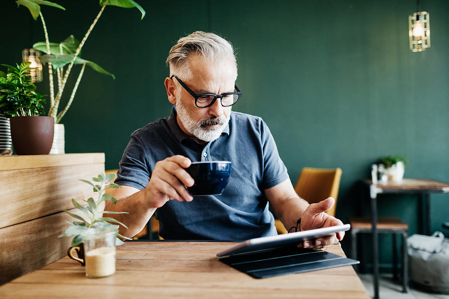 Mature Man Sitting In Cafe Reading On Tablet Photograph by Tom Werner