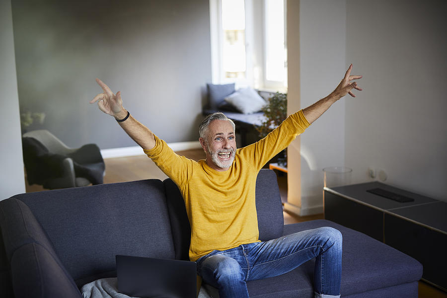 Mature man sitting on couch at home cheering Photograph by Westend61
