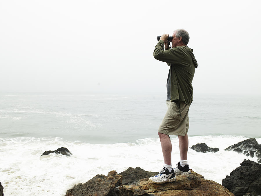 Mature man with binoculars standing on rocks on beach Photograph by Ryan McVay