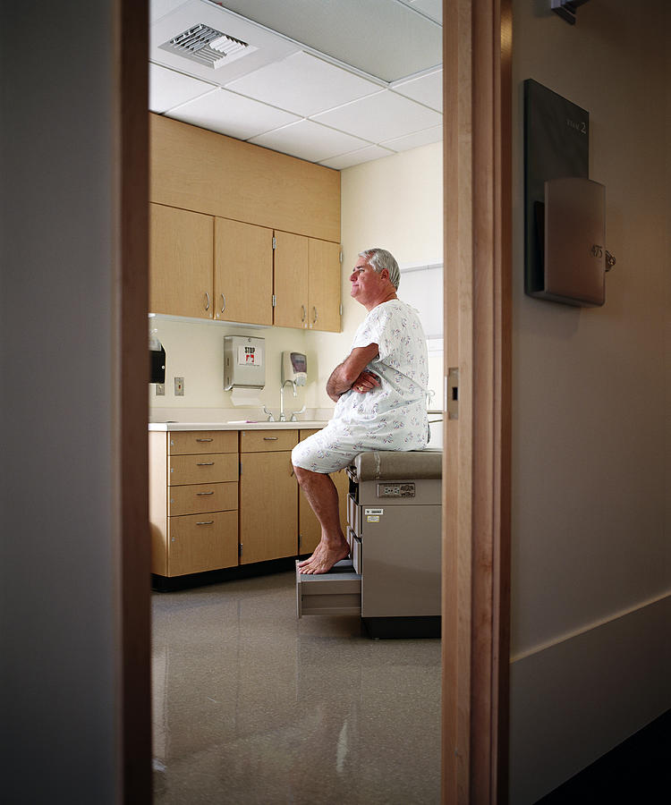 Mature patient wearing hospital gown, sitting on examing table Photograph by Thomas Barwick