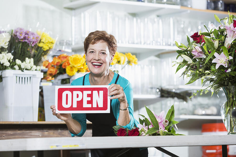 Mature woman in flower shop holding OPEN sign Photograph by Kali9