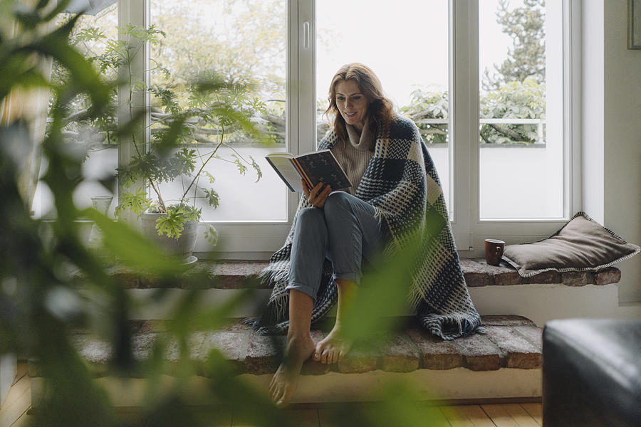 Mature woman sitting on wondow sill, wrapped in blanket, reading book Photograph by Westend61