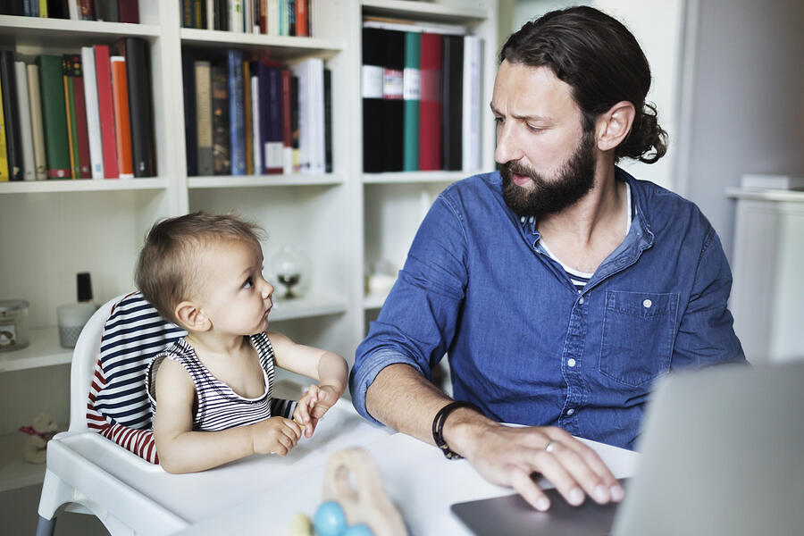 Mid adult businessman using laptop while looking at baby boy at home Photograph by Maskot