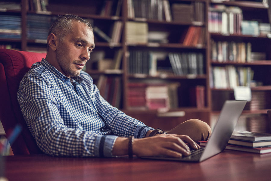 Mid adult businessman working on laptop at home library. Photograph by Skynesher