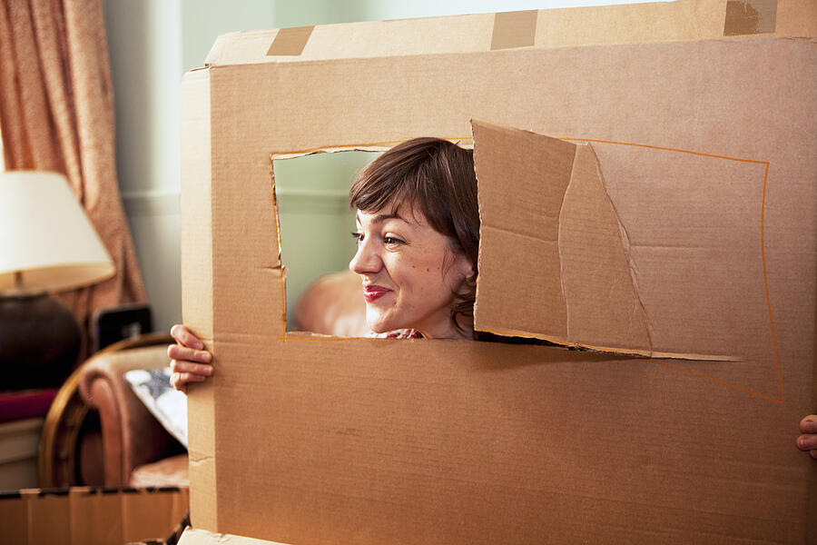Mid adult woman peeking out of cardboard box window in living room Photograph by Nancy Honey