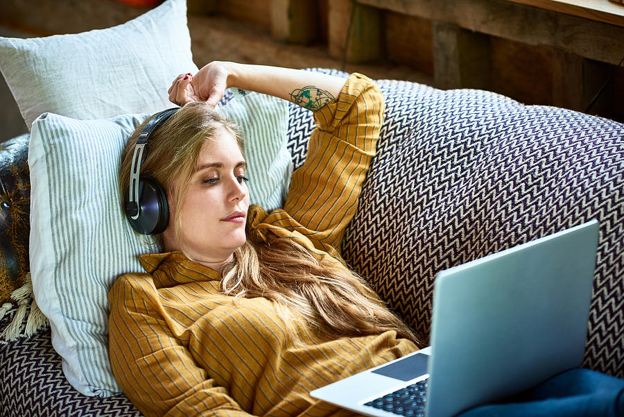 Mid adult woman reclining on sofa with laptop wearing headphones Photograph by 10000 Hours